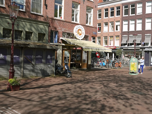 A street scene featuring a red-brick building housing a shop labeled 'Bagels & Beans' with a cream-colored awning. In front of the shop, there is a row of bicycles parked on the sidewalk. A person in a blue outfit is walking with a bag. A vintage-style lamppost is situated on the left, and shadows of trees are cast on the ground.