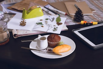 A cozy workspace scene with a table covered in various craft and food items. A white plate holds a chocolate muffin, a small creamer, and a pastry, next to a fork and a knife. Nearby, there's a glass mug and a digital tablet. Papers and notebooks are scattered, with one sheet displaying dried leaves and plants. Crafting supplies like scissors, a glue stick, and a lime green tape dispenser complete the scene.