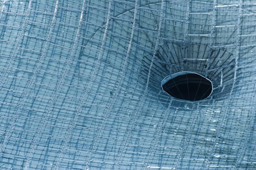 A complex network of metallic grid structures forms a curved funnel shape with a dark circular opening at its center. The mesh-like pattern is symmetrical and uniform, reflecting light and creating a futuristic and technical appearance.