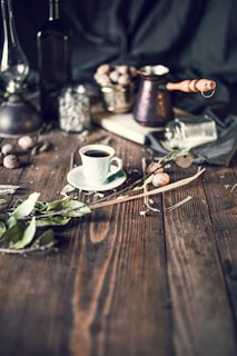 An artistic arrangement of a rustic kitchen setting featuring a cup of coffee on a saucer, surrounded by various objects including a cezve (Turkish coffee pot), a jar, and scattered leaves. Walnuts in a metal container add a natural touch, while a dark, moody background contrasts with the warm, earthy tones of the wooden surface.