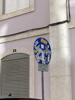 A traffic sign covered with various stickers stands in front of a light-colored building wall. The sign has a smaller sign beneath it reading 'EXCEPTO TÁXIS'. There is also an arched, closed shutter nearby, and a building number '39' above. Electrical cables run horizontally across the wall above the sign.