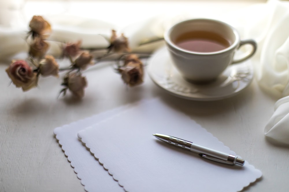 tasse en céramique blanche sur soucoupe en céramique blanche à côté du stylo à clic argenté