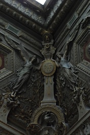 Intricate architectural details featuring two sculpted figures with wings flanking a central medallion inscribed with 'R.F'. The decor includes ornate foliage, geometric patterns, and a bird motif near the base. The artwork is in a rich, dark palette, indicating a sense of antiquity and elegance.