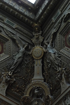 Intricate architectural details featuring two sculpted figures with wings flanking a central medallion inscribed with 'R.F'. The decor includes ornate foliage, geometric patterns, and a bird motif near the base. The artwork is in a rich, dark palette, indicating a sense of antiquity and elegance.