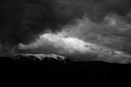 Dark storm clouds loom over a rugged mountain range, partially covered with snow, creating a dramatic and intense atmosphere. A patch of lighter clouds breaks through, highlighting the mountain peaks below.