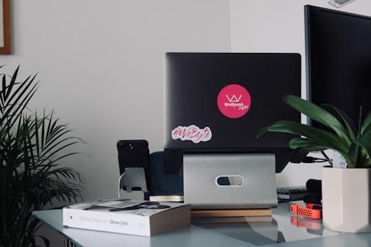 A modern workspace featuring a laptop with stickers, a book titled 'Steve Jobs' by Walter Isaacson, and various pieces of office equipment. Green plants add a touch of nature, and there is a mix of technology and personal items.