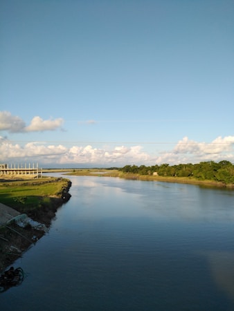 A serene river flows smoothly through the landscape, bordered by grassy banks and dense greenery. In the distance, a construction site with an unfinished building framework is visible. The sky is clear with a few scattered clouds, creating a peaceful ambiance.