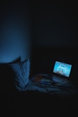 A dimly lit room with a laptop on a bed, displaying a Paramount Pictures logo. The room is dark with a faint blue glow from the laptop screen illuminating the surroundings. A pillow rests on the bed next to the laptop.