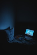 A dimly lit room with a laptop on a bed, displaying a Paramount Pictures logo. The room is dark with a faint blue glow from the laptop screen illuminating the surroundings. A pillow rests on the bed next to the laptop.
