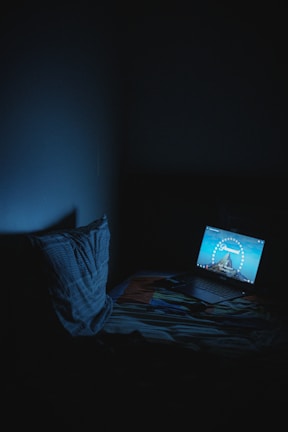 A dimly lit room with a laptop on a bed, displaying a Paramount Pictures logo. The room is dark with a faint blue glow from the laptop screen illuminating the surroundings. A pillow rests on the bed next to the laptop.