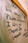 The image displays a message written on a glass or whiteboard with a wooden frame. The message reads 'Dear me, I'm sorry, Please forgive me, I love you, Thank you' with further instructions to repeat it. There is also a small decorative symbol on the right side.
