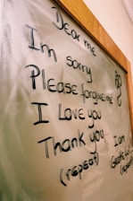 The image displays a message written on a glass or whiteboard with a wooden frame. The message reads 'Dear me, I'm sorry, Please forgive me, I love you, Thank you' with further instructions to repeat it. There is also a small decorative symbol on the right side.
