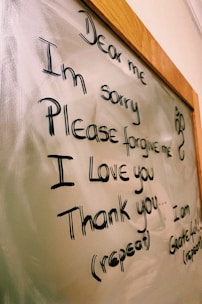The image displays a message written on a glass or whiteboard with a wooden frame. The message reads 'Dear me, I'm sorry, Please forgive me, I love you, Thank you' with further instructions to repeat it. There is also a small decorative symbol on the right side.