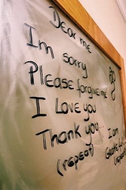 The image displays a message written on a glass or whiteboard with a wooden frame. The message reads 'Dear me, I'm sorry, Please forgive me, I love you, Thank you' with further instructions to repeat it. There is also a small decorative symbol on the right side.