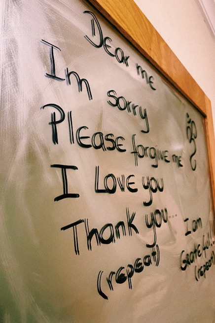 The image displays a message written on a glass or whiteboard with a wooden frame. The message reads 'Dear me, I'm sorry, Please forgive me, I love you, Thank you' with further instructions to repeat it. There is also a small decorative symbol on the right side.
