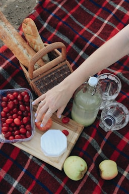 A cozy picnic setup on a red and black checkered blanket with a variety of items including a basket with baguettes, a cutting board with a round piece of cheese and a sliced apple, a container of cherry tomatoes, a bottle of a light-colored drink, two clear glasses, and a woman's hand reaching towards the board. Two apples are placed nearby.