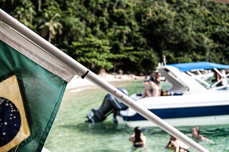 A Brazilian flag is prominently displayed in the foreground, with a vibrant green and yellow color scheme. In the background, a group of people enjoy swimming in clear, shallow waters near a large white and blue motorboat. The setting appears tropical, with lush green foliage and a sandy beach visible in the distance.