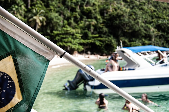 A Brazilian flag is prominently displayed in the foreground, with a vibrant green and yellow color scheme. In the background, a group of people enjoy swimming in clear, shallow waters near a large white and blue motorboat. The setting appears tropical, with lush green foliage and a sandy beach visible in the distance.