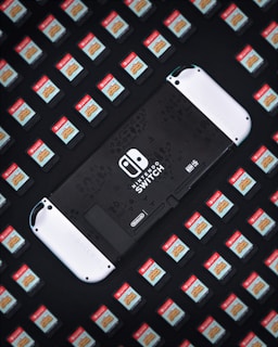 A Nintendo Switch console is surrounded by numerous game cartridges, each featuring a red label with a game title. The console is positioned in the center, partially visible, and is predominantly black with white accents.