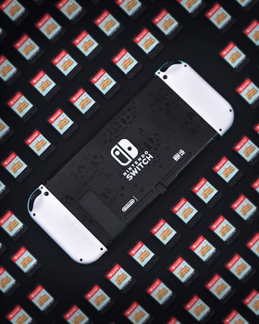 A Nintendo Switch console is surrounded by numerous game cartridges, each featuring a red label with a game title. The console is positioned in the center, partially visible, and is predominantly black with white accents.