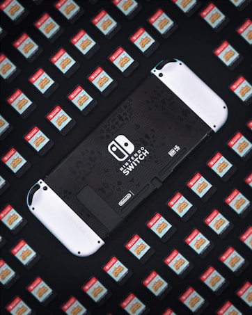 A Nintendo Switch console is surrounded by numerous game cartridges, each featuring a red label with a game title. The console is positioned in the center, partially visible, and is predominantly black with white accents.