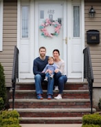 A family of three is sitting on the steps in front of a house. The father, mother, and child are dressed casually. A pink floral wreath decorates the front door, which has the house number 688 displayed. The entrance is flanked by black iron railings and the front porch is surrounded by small bushes.