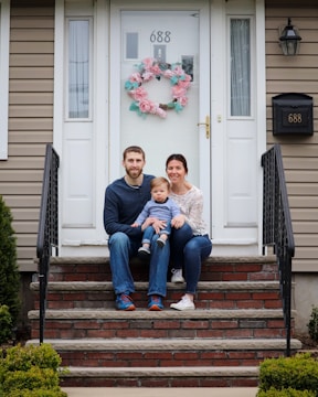 A family of three is sitting on the steps in front of a house. The father, mother, and child are dressed casually. A pink floral wreath decorates the front door, which has the house number 688 displayed. The entrance is flanked by black iron railings and the front porch is surrounded by small bushes.