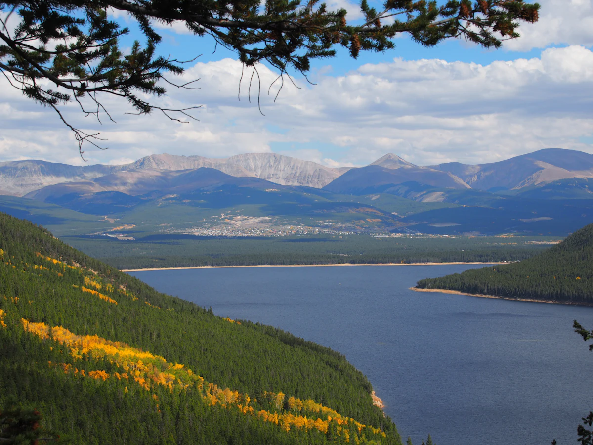 A lake below forested slopes with yellow aspens near Leadville, with a town and a mountain range beyond