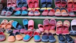 A variety of colorful, handcrafted shoes arranged in neat rows. The shoes display intricate patterns and vibrant colors such as red, blue, orange, and black. Some feature detailed embroidery or unique designs, highlighting artisanal craftsmanship.