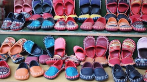 A variety of colorful, handcrafted shoes arranged in neat rows. The shoes display intricate patterns and vibrant colors such as red, blue, orange, and black. Some feature detailed embroidery or unique designs, highlighting artisanal craftsmanship.