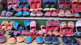 A variety of colorful, handcrafted shoes arranged in neat rows. The shoes display intricate patterns and vibrant colors such as red, blue, orange, and black. Some feature detailed embroidery or unique designs, highlighting artisanal craftsmanship.