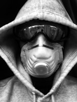 A person wearing a hoodie, goggles, and a respiratory mask covers their face entirely, creating a mysterious and protective appearance. The image is in black and white.