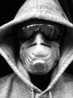 A person wearing a hoodie, goggles, and a respiratory mask covers their face entirely, creating a mysterious and protective appearance. The image is in black and white.