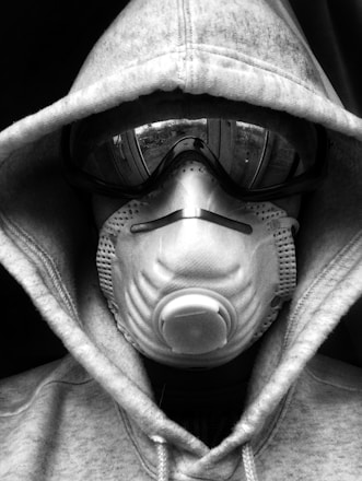 A person wearing a hoodie, goggles, and a respiratory mask covers their face entirely, creating a mysterious and protective appearance. The image is in black and white.