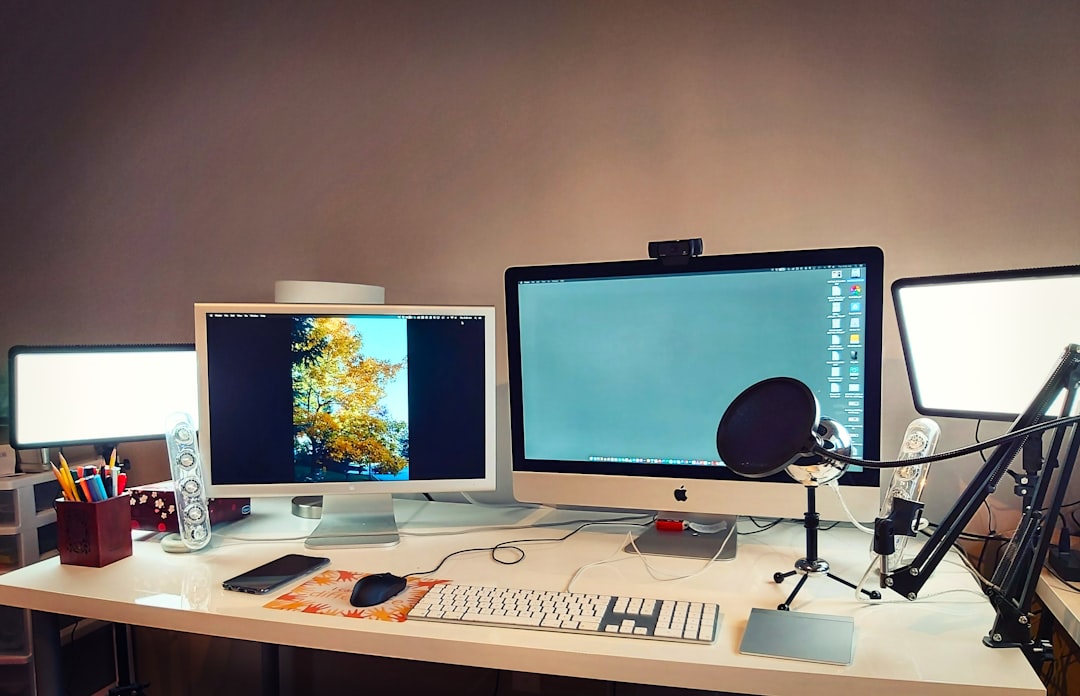 silver imac on brown wooden desk, Modern Home Office Setup With Dual Monitors, Desk Accessories, and Task Lighting in Evening Hours - A well-organized home office features two large monitors displaying vibrant images and a clean workspace. The desk is adorned with various stationery items, a microphone, and ambient task lighting, creating a productive atmosphere during evening hours.