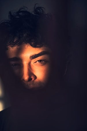 A mysterious portrait showcasing a person's face partially illuminated by warm, dramatic lighting, creating deep shadows. The individual's features are accentuated, with one eye prominently catching the light, adding intrigue.