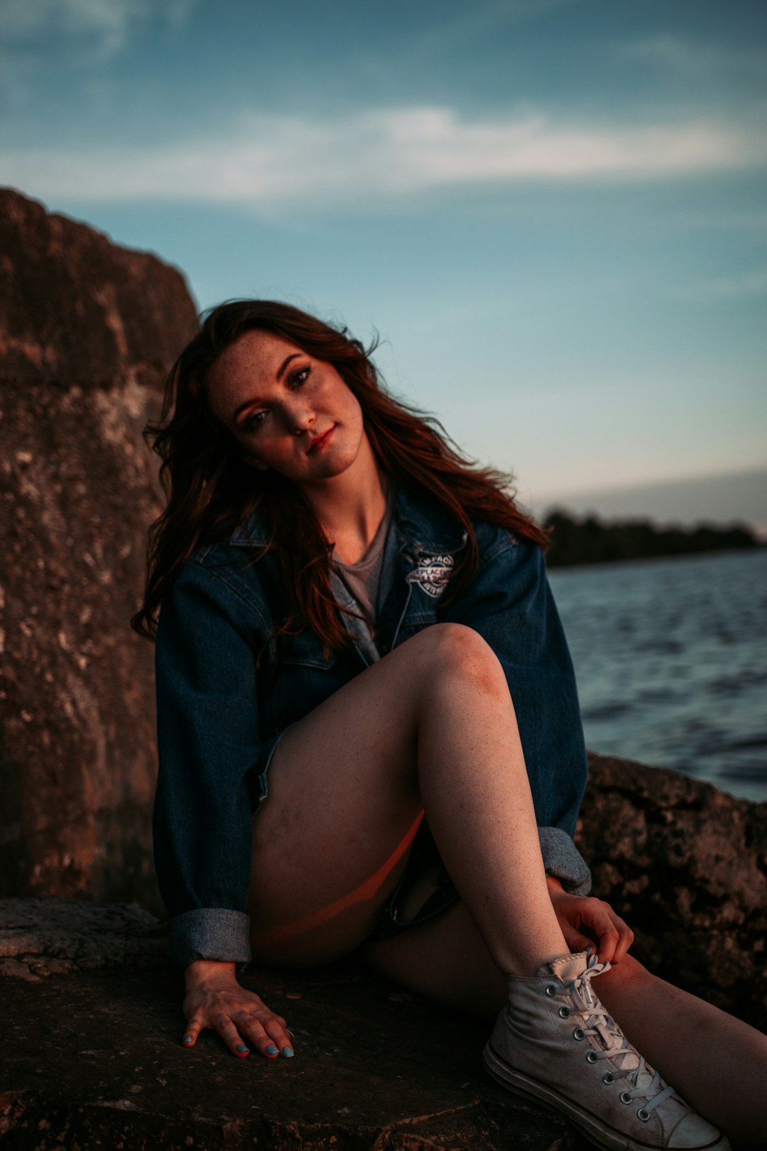 woman in blue denim jacket sitting on rock near body of water during daytime