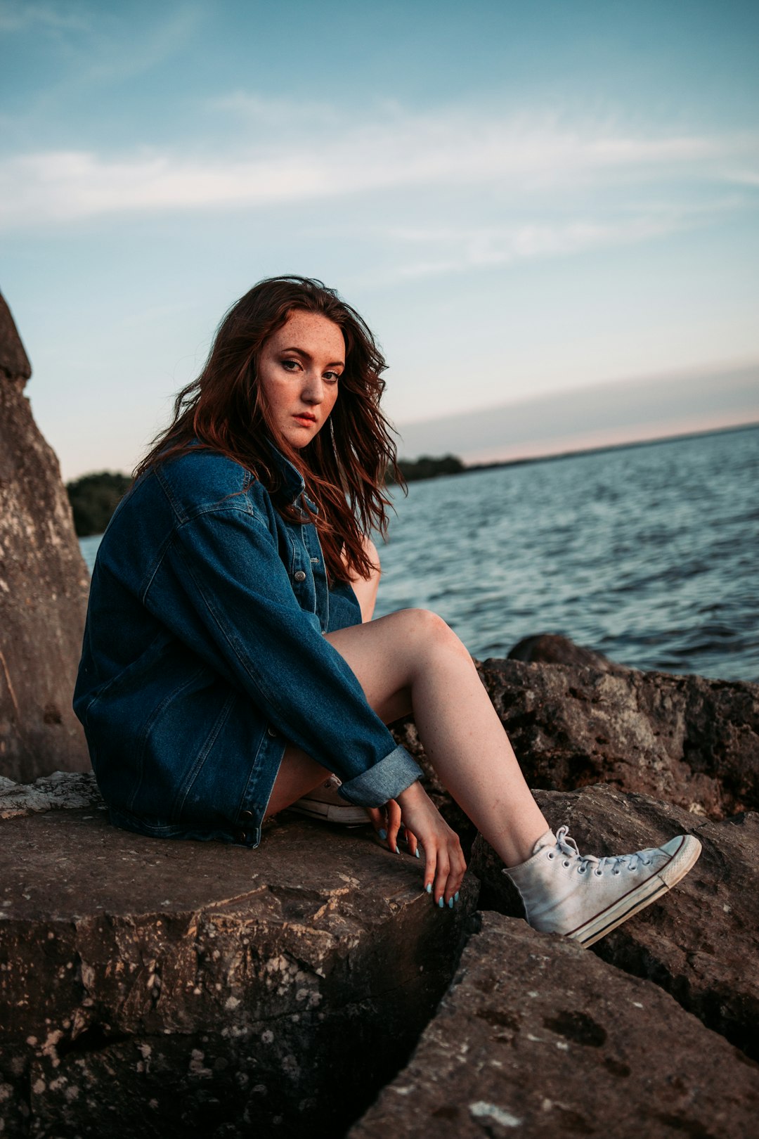 woman in blue denim jacket sitting on brown rock near body of water during daytime