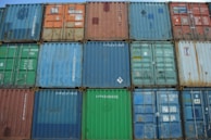 A stacked arrangement of colorful shipping containers, each marked with various numbers and logos. The containers display signs of wear and rust, indicating frequent use, and are arranged in a tight grid pattern.