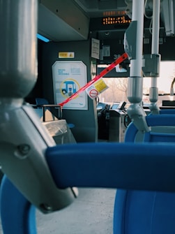 The interior of a public bus with blue seats is visible, featuring a barrier with red tape and a warning sign near the front section. A digital display is present at the top with illuminated text. The overall scene appears quiet and unoccupied, with metal poles and handles for passengers.