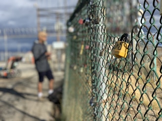 A chain-link fence with several locks attached, focusing primarily on a yellow lock in the foreground. In the background, a blurry figure is standing next to machinery, and there are signs and utility structures.
