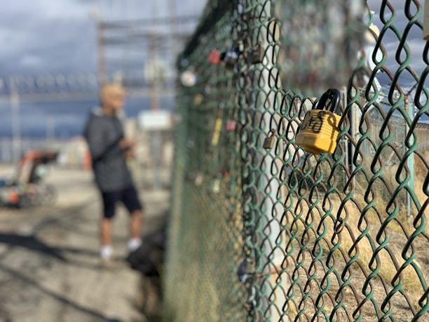 A chain-link fence with several locks attached, focusing primarily on a yellow lock in the foreground. In the background, a blurry figure is standing next to machinery, and there are signs and utility structures.