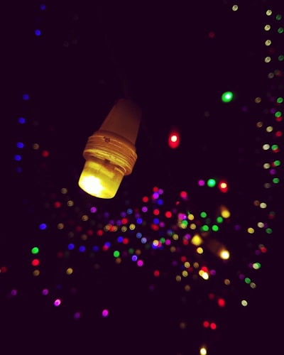 A close-up of a glowing yellow LED bulb against a dark background, with numerous blurred colorful lights in the distance, creating a bokeh effect.