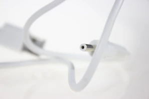 A close-up view of a white cable with a cylindrical connector in focus, set against a blurred white background. The cable curves gracefully, leading the eye to the connector tip.