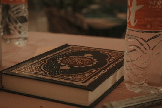 A table is set with a decorative book with intricate patterns on its cover, placed beside a partially visible plastic bottle of water and a pen. The ambiance suggests a calm and meditative environment possibly for study or reading.