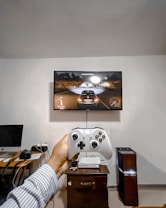 A person is holding a gaming controller in front of a flat-screen television displaying a racing video game. The room has a computer setup on a wooden desk with cables, a desktop computer monitor, and other accessories. The focus is on the interaction between the person and the gaming environment.