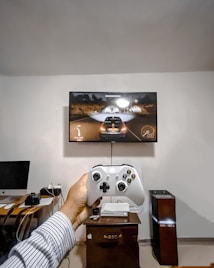 A person is holding a gaming controller in front of a flat-screen television displaying a racing video game. The room has a computer setup on a wooden desk with cables, a desktop computer monitor, and other accessories. The focus is on the interaction between the person and the gaming environment.