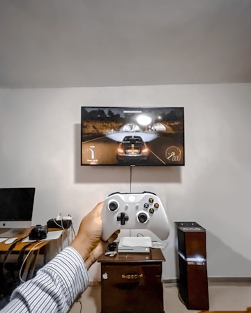 A person is holding a gaming controller in front of a flat-screen television displaying a racing video game. The room has a computer setup on a wooden desk with cables, a desktop computer monitor, and other accessories. The focus is on the interaction between the person and the gaming environment.