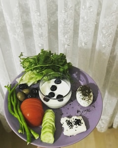 A purple plate holds a variety of fresh and healthy foods. There are long green beans, black and green olives, a sliced tomato, sliced cucumber, a bunch of leafy greens, and a glass filled with yogurt topped with blueberries. A boiled egg is garnished with herbs, and a small square of cheese is sprinkled with black seeds.