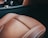 A close-up view of the interior of a luxury car, showcasing a brown leather seat with light stitching and perforated details. The background includes part of the car's dashboard and a metallic control knob, with accents of black and carbon fiber texture.